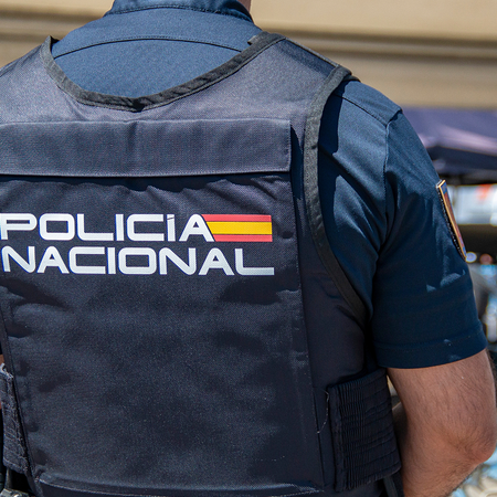 A Policía Nacional officer wearing a protective vest marked "Policía Nacional" stands with his back to the camera at an outdoor event.