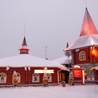 Snow-covered red wooden buildings decorated with Christmas lights at Santa Claus Village in Rovaniemi, Finland, with a "Meet Santa" sign out front.
