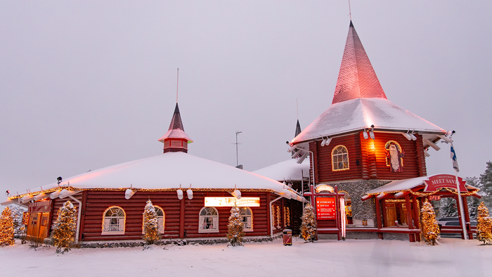 Snow-covered red wooden buildings decorated with Christmas lights at Santa Claus Village in Rovaniemi, Finland, with a "Meet Santa" sign out front.