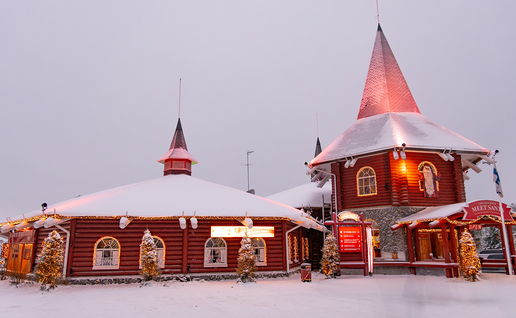 Snow-covered red wooden buildings decorated with Christmas lights at Santa Claus Village in Rovaniemi, Finland, with a "Meet Santa" sign out front.