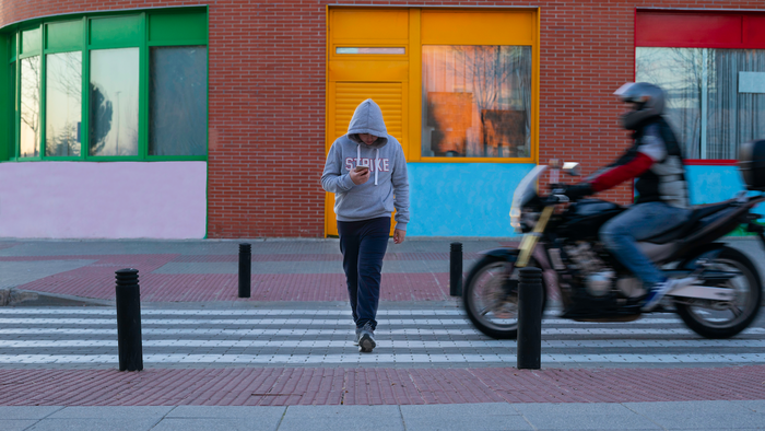 A man crossing a zebra crossing wearing a grey hoodie looking at his phone with a motorbike almost hitting him