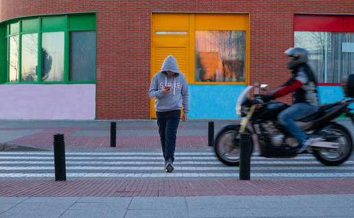 A man crossing a zebra crossing wearing a grey hoodie looking at his phone with a motorbike almost hitting him