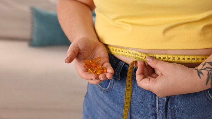 Woman in a yellow shirt measures her waist with a tape measure whilst holding slimming pills