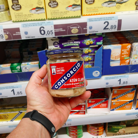 A mans hand holding a jar of tuna with different cans are stocked on the shelf behind