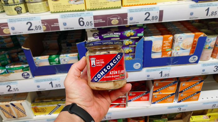 A mans hand holding a jar of tuna with different cans are stocked on the shelf behind
