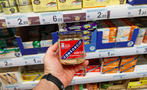 A mans hand holding a jar of tuna with different cans are stocked on the shelf behind