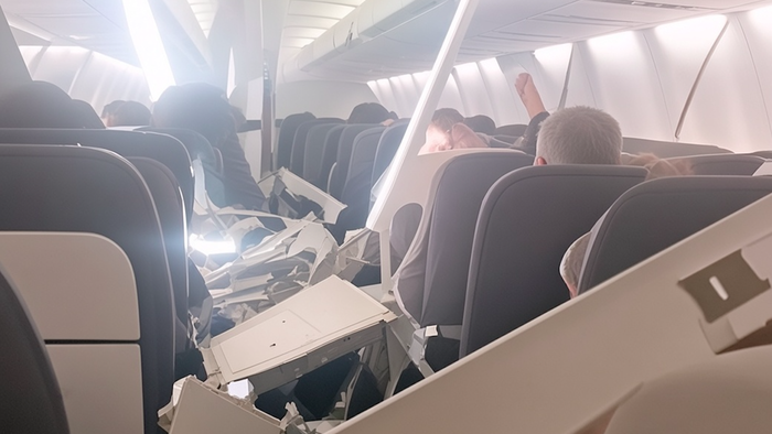 Airplane cabin interior with collapsed white plastic ceiling panels resting on seats and passengers.