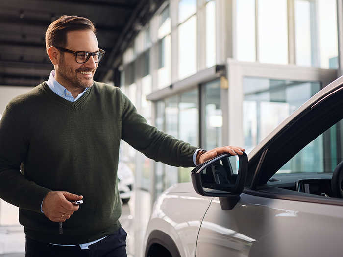A man smiling as he opens the door of a new car in a dealership showroom, holding the keys.