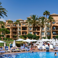 Guests relax on sun loungers by a hotel swimming pool, with palm trees and apartment-style hotel blocks in the background.