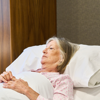 An elderly woman lying in bed, looking away from the camera, hands resting on the covers