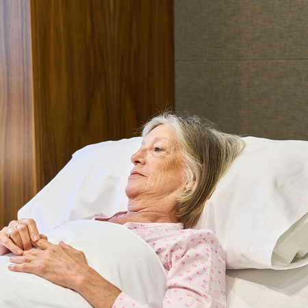 An elderly woman lying in bed, looking away from the camera, hands resting on the covers