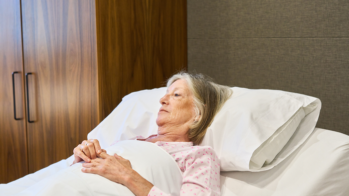 An elderly woman lying in bed, looking away from the camera, hands resting on the covers