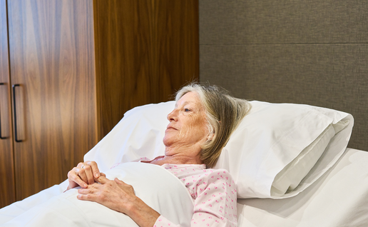 An elderly woman lying in bed, looking away from the camera, hands resting on the covers