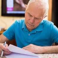 An elderly man sits at a table, reading and writing on a paper document.