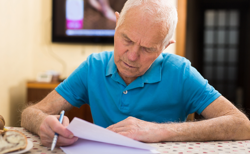 An elderly man sits at a table, reading and writing on a paper document.