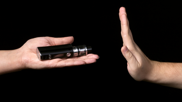 A hand offers a black vape device while another hand held up in a stop gesture refuses it, against a plain black background.