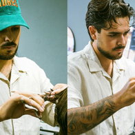 A professional male barber with tattooed arms using scissors and a comb to trim a client's hair in a barbershop setting.