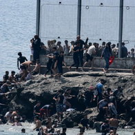 Large group of migrants climb rocks and enter Ceuta from Morocco beside the border fence during the July 2026 invasion.