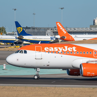 An orange and white easyJet plane taxiing on an airport runway, with two Ryanair jets and another easyJet plane parked behind it under a clear blue sky.