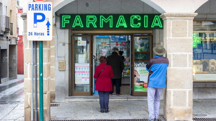 Senior patients waiting outside a Spanish pharmacy ("Farmacia") storefront with a green glowing sign, illustrating prescription management and community health care.