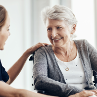 A carer sits with an elderly woman in a wheelchair as they talk indoors.