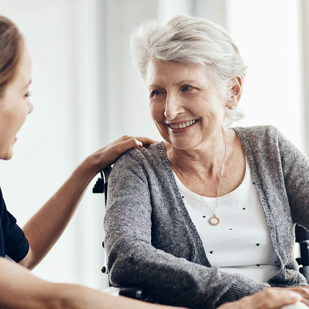 A carer sits with an elderly woman in a wheelchair as they talk indoors.