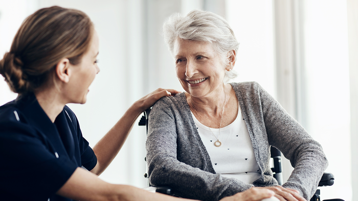 A carer sits with an elderly woman in a wheelchair as they talk indoors.