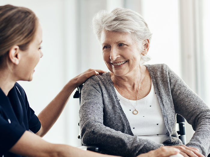 A carer sits with an elderly woman in a wheelchair as they talk indoors.