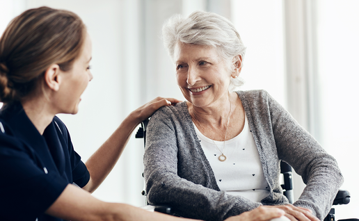 A carer sits with an elderly woman in a wheelchair as they talk indoors.