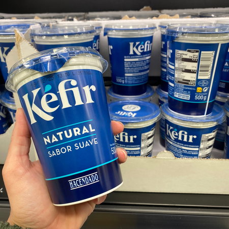 A hand holding a Kefir yogurt with others in the refrigerator in the background