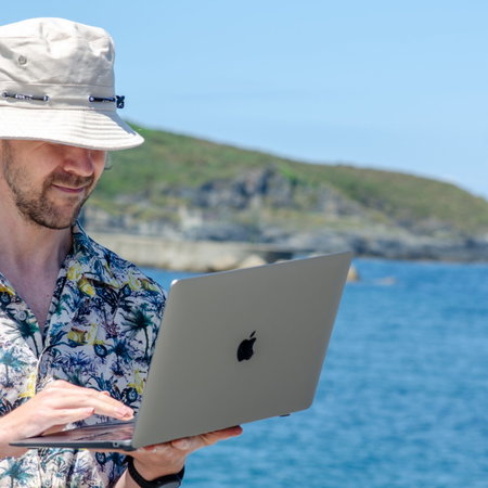 A man in a Hawaiian shirt wearing a bucket hat and holding an open laptop next to the ocean