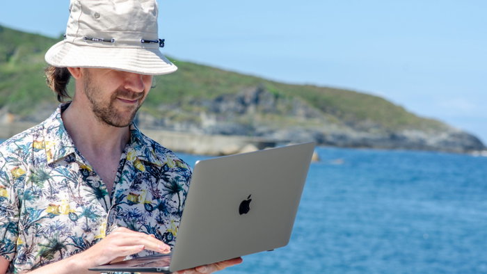 A man in a Hawaiian shirt wearing a bucket hat and holding an open laptop next to the ocean
