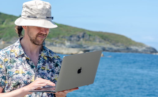 A man in a Hawaiian shirt wearing a bucket hat and holding an open laptop next to the ocean