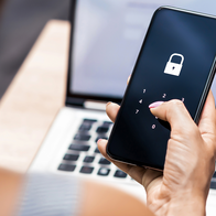 Woman’s hand entering a passcode on a smartphone screen showing a padlock icon, with a laptop open behind her.