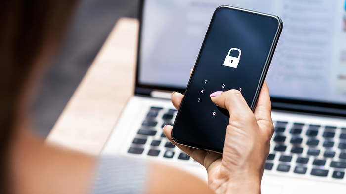 Woman’s hand entering a passcode on a smartphone screen showing a padlock icon, with a laptop open behind her.
