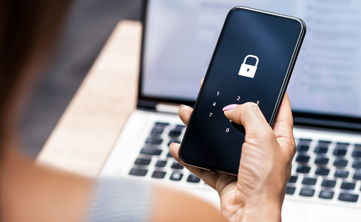 Woman’s hand entering a passcode on a smartphone screen showing a padlock icon, with a laptop open behind her.