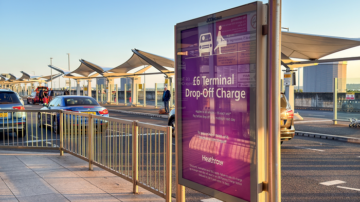 A purple Heathrow sign by a terminal drop-off lane reading “£6 Terminal Drop-Off Charge”, with cars and passengers under canopies in the background