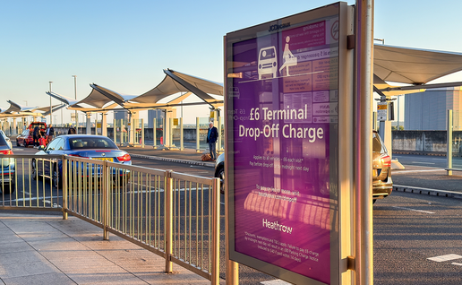 A purple Heathrow sign by a terminal drop-off lane reading “£6 Terminal Drop-Off Charge”, with cars and passengers under canopies in the background