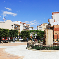 A sunny paved town square in Albox, with a stone fountain inside a low iron railing, trimmed trees, benches and flat-fronted apartment buildings under a deep blue sky.
