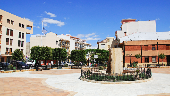 A sunny paved town square in Albox, with a stone fountain inside a low iron railing, trimmed trees, benches and flat-fronted apartment buildings under a deep blue sky.
