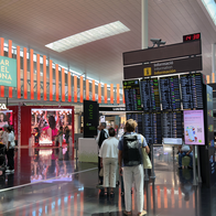 Travellers checking departure boards under an information sign in a busy airport terminal, with duty-free shops and seating around them.