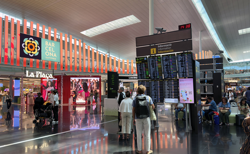 Travellers checking departure boards under an information sign in a busy airport terminal, with duty-free shops and seating around them.