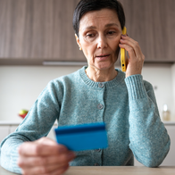 Woman with short dark hair in a blue-green cardigan holds a bank card while on a phone call at a kitchen table.