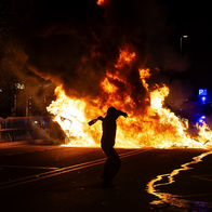 A hooded figure in silhouette swings an arm back to throw a bottle towards a large street fire at night, while another person photographs the flames and police vehicles with flashing blue lights stand in the background.