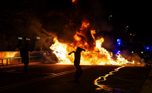 A hooded figure in silhouette swings an arm back to throw a bottle towards a large street fire at night, while another person photographs the flames and police vehicles with flashing blue lights stand in the background.