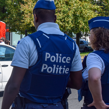 Two Belgian police officers in blue caps and body armour marked “Police” and “Politie” stand with their backs to the camera beside a white police car, with a crowd and a stage under a red and green canopy behind them.