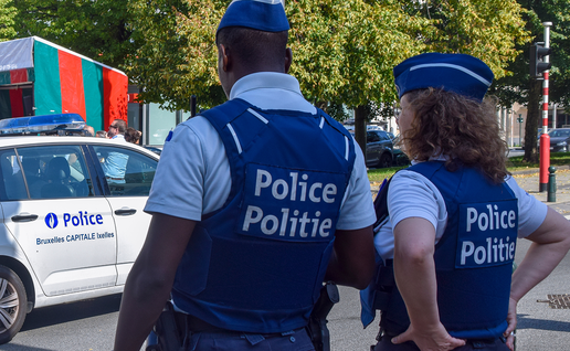 Two Belgian police officers in blue caps and body armour marked “Police” and “Politie” stand with their backs to the camera beside a white police car, with a crowd and a stage under a red and green canopy behind them.