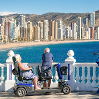 An older couple at a white seafront viewpoint looking out over a curved sandy beach and blue sea. The woman sits on a mobility scooter and the man leans on the railing, with a row of high-rise towers and dry hills behind the beach.