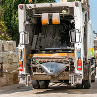 The back of a white bin lorry with its rear loading area open, parked on a paved street between an old stone wall and a building, with a brick bell tower and blocks of flats in the background.