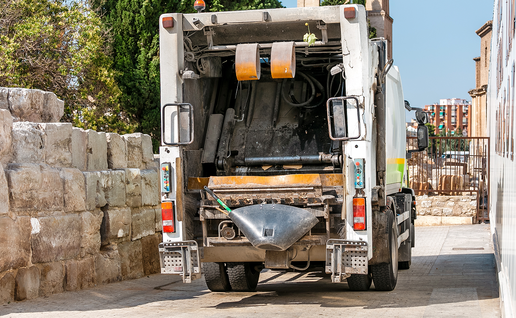The back of a white bin lorry with its rear loading area open, parked on a paved street between an old stone wall and a building, with a brick bell tower and blocks of flats in the background.